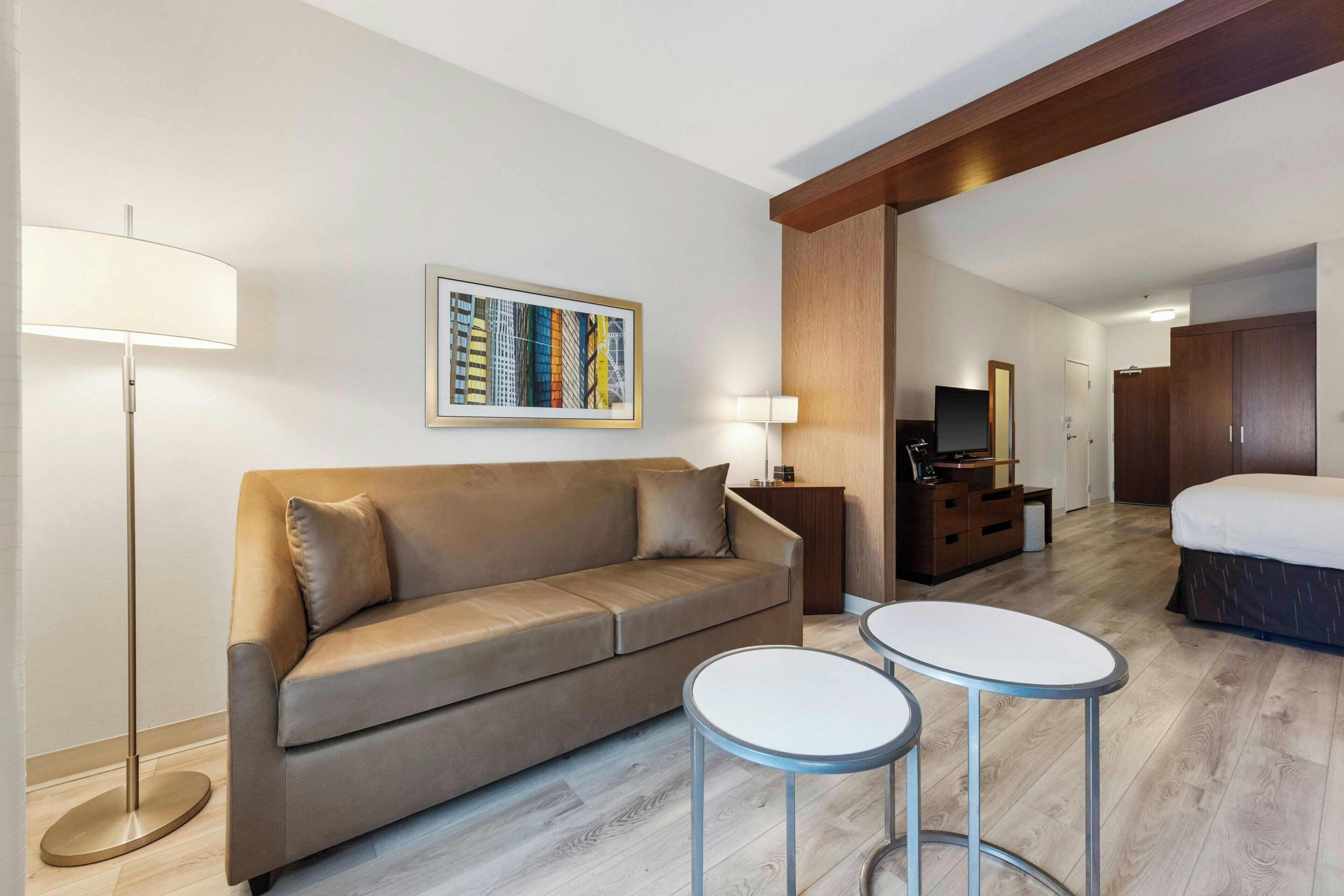 A hotel room with a sofa, two small tables, a lamp, a TV, a wardrobe, and a painting on the wall.
