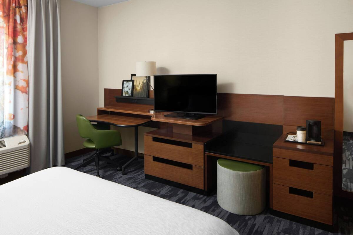 A hotel room with a bed, a desk with a television, a green chair, a waste bin, and a lamp.
