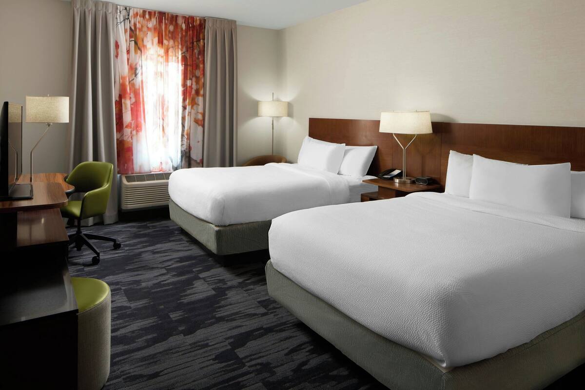 A hotel room with two beds, a desk with a chair, a lamp, and a large window with curtains.