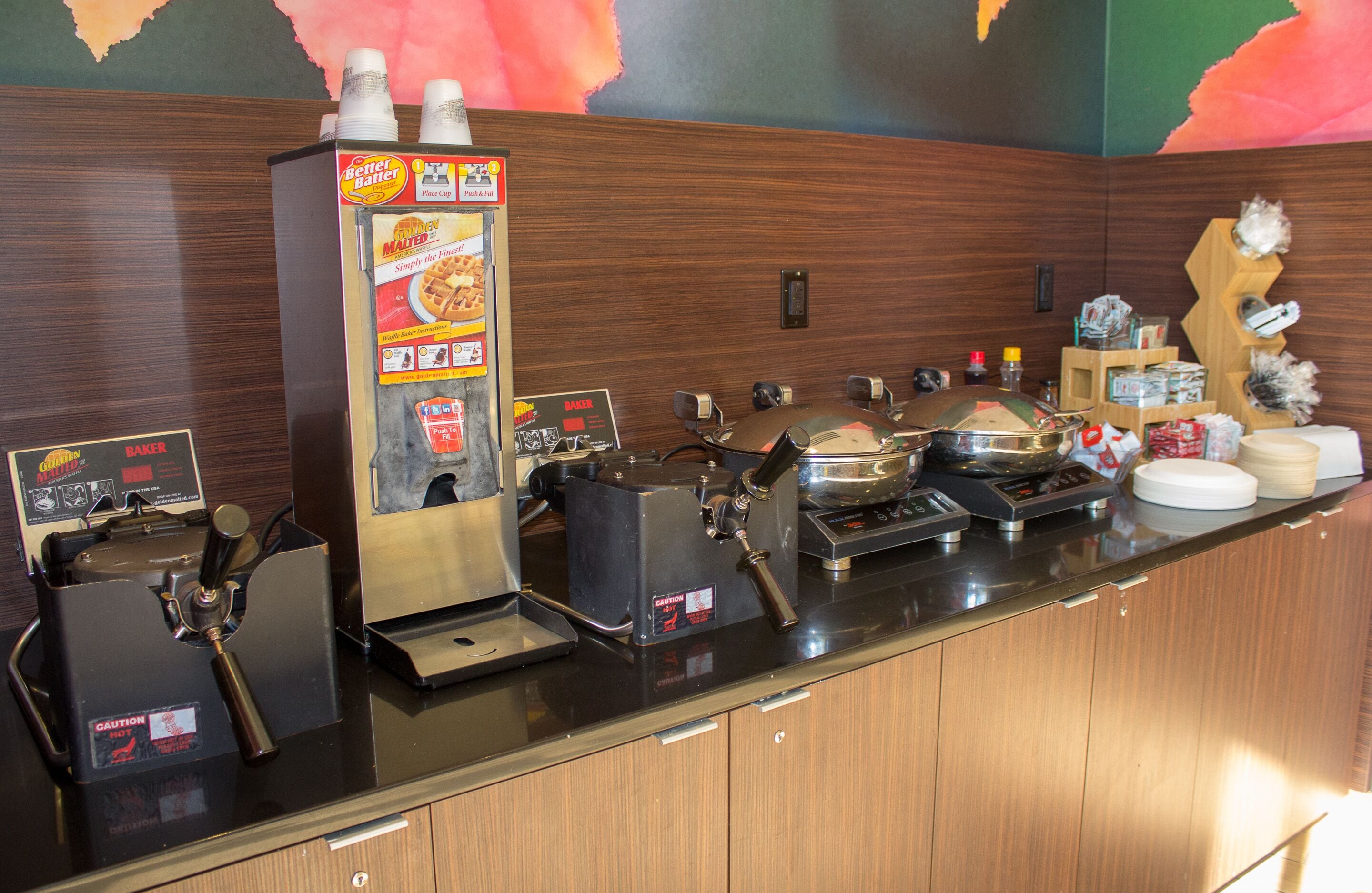A self-service breakfast station with coffee machines, a waffle maker, and various condiments.