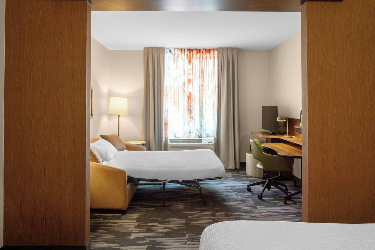 A hotel room with a bed, a desk, a chair, a lamp, and a window with curtains.