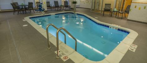 Una piscina interior con una escalera, sillas y suelo de baldosas.