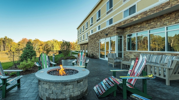 A stone fire pit surrounded by Adirondack chairs and a seating area with skis and snowboards leaning against a wall. - Fairfield by Marriott Inn & Suites Plymouth White Mountains (Plymouth)