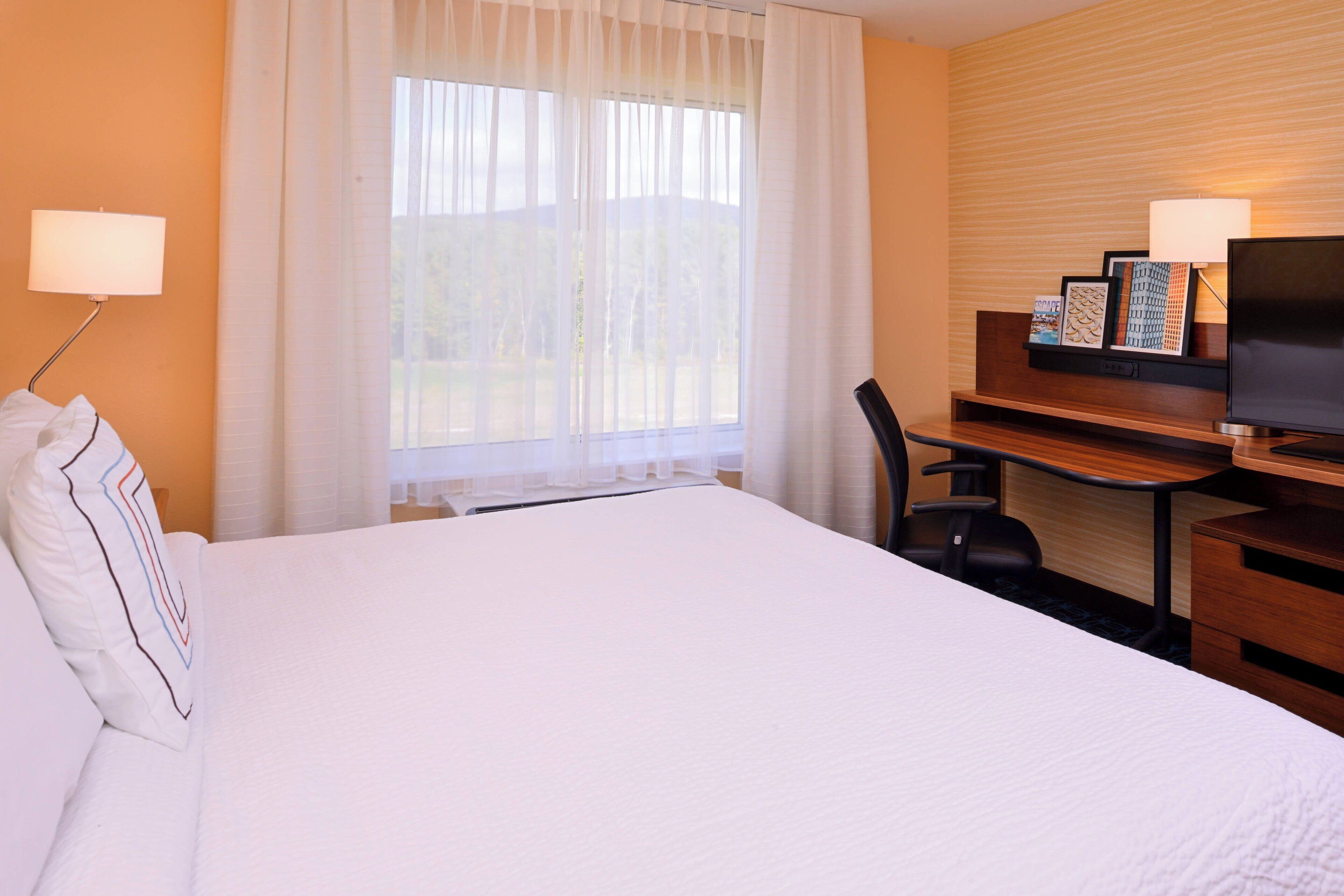 A hotel room with a large bed, a desk with a chair, a television, and a window with sheer curtains.