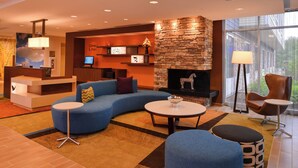 A modern hotel lobby with a curved blue sofa, a stone fireplace, a reception desk, and a large window offering a view of the outdoors. - Fairfield by Marriott Inn & Suites Plymouth White Mountains (Plymouth)