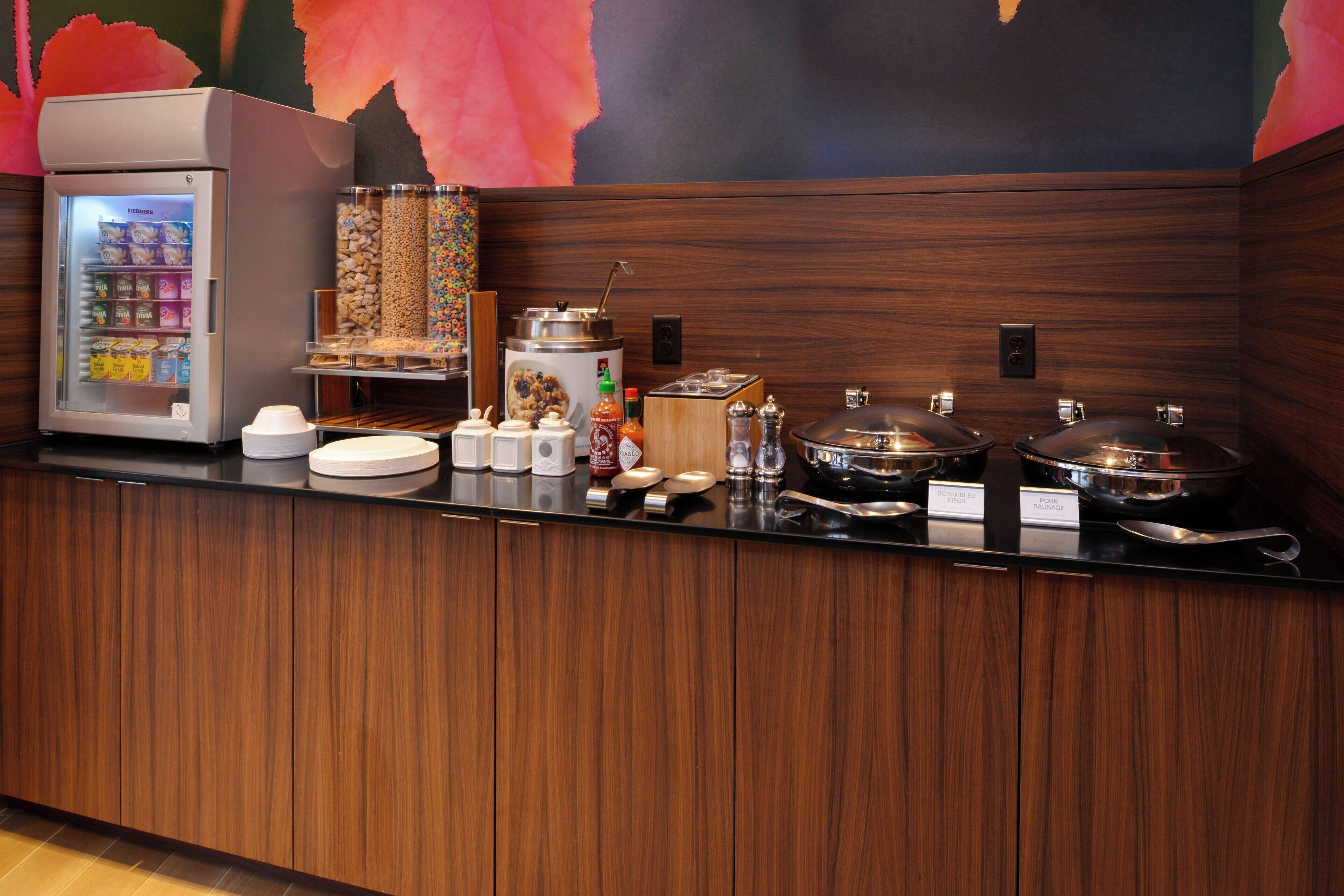 A hotel reception area with a self-service beverage station, including a refrigerated drink cooler, various condiments, and a coffee machine.
