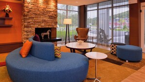 A modern living room with a stone fireplace, a blue sofa, a round coffee table, and a view of the outdoors through large windows. - Fairfield by Marriott Inn & Suites Plymouth White Mountains (Plymouth)