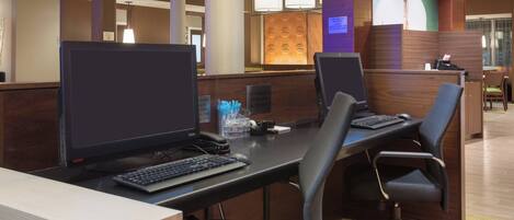 A hotel reception area with a wooden reception desk, two computer workstations, and a seating area with chairs.
