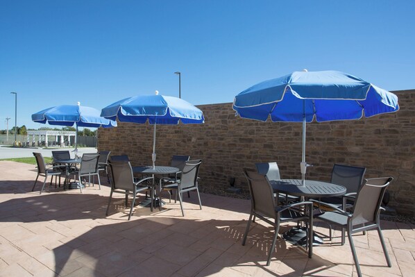 Un patio exterior con paredes de piedra, mesas y sillas de metal, y sombrillas azules que dan sombra.