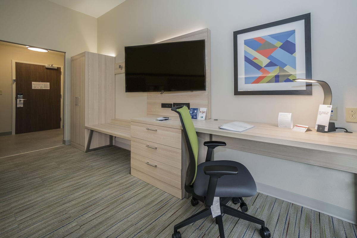 A hotel room with a desk, chair, TV, artwork, and a door.
