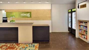 A hotel reception area with a reception desk, a sign reading "HOME 2 SUITES BY HILTON," and a refrigerator stocked with beverages.