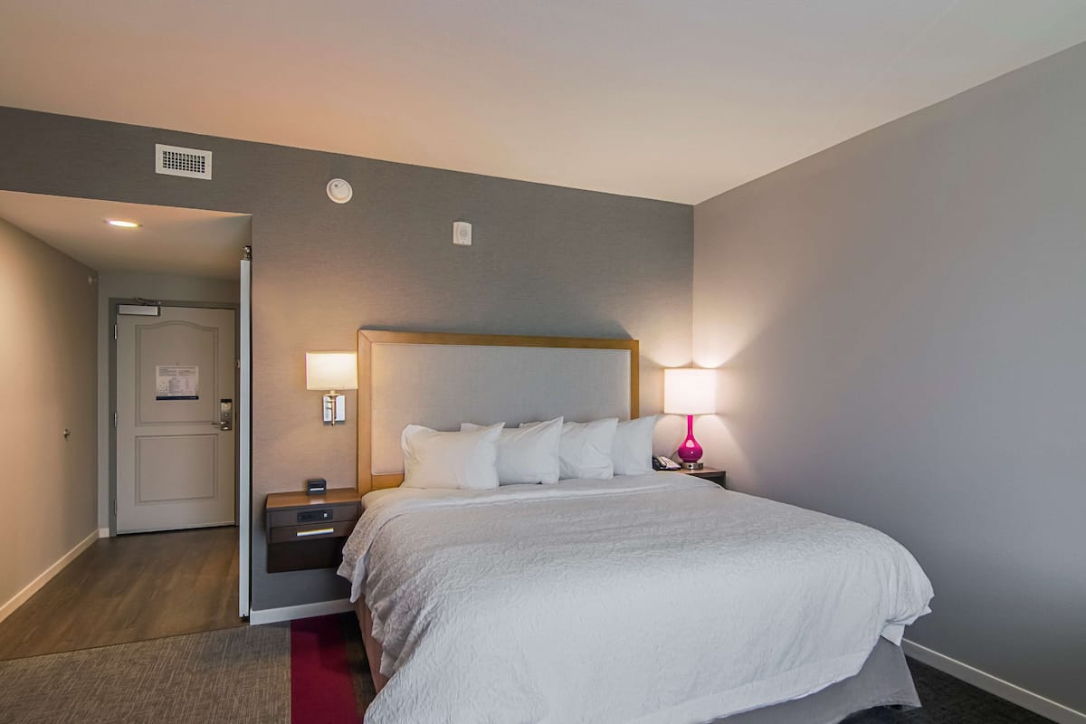 Habitación de hotel con una cama grande, una mesita de noche, una lámpara y una puerta que da a otra habitación.