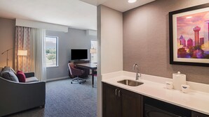Quarto de hotel com cozinha compacta, área de estar com sofá e poltrona, televisão e uma janela grande com cortinas.