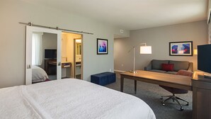 Quarto de hotel com uma cama grande, uma escrivaninha, uma cadeira, um sofá e duas obras de arte nas paredes.