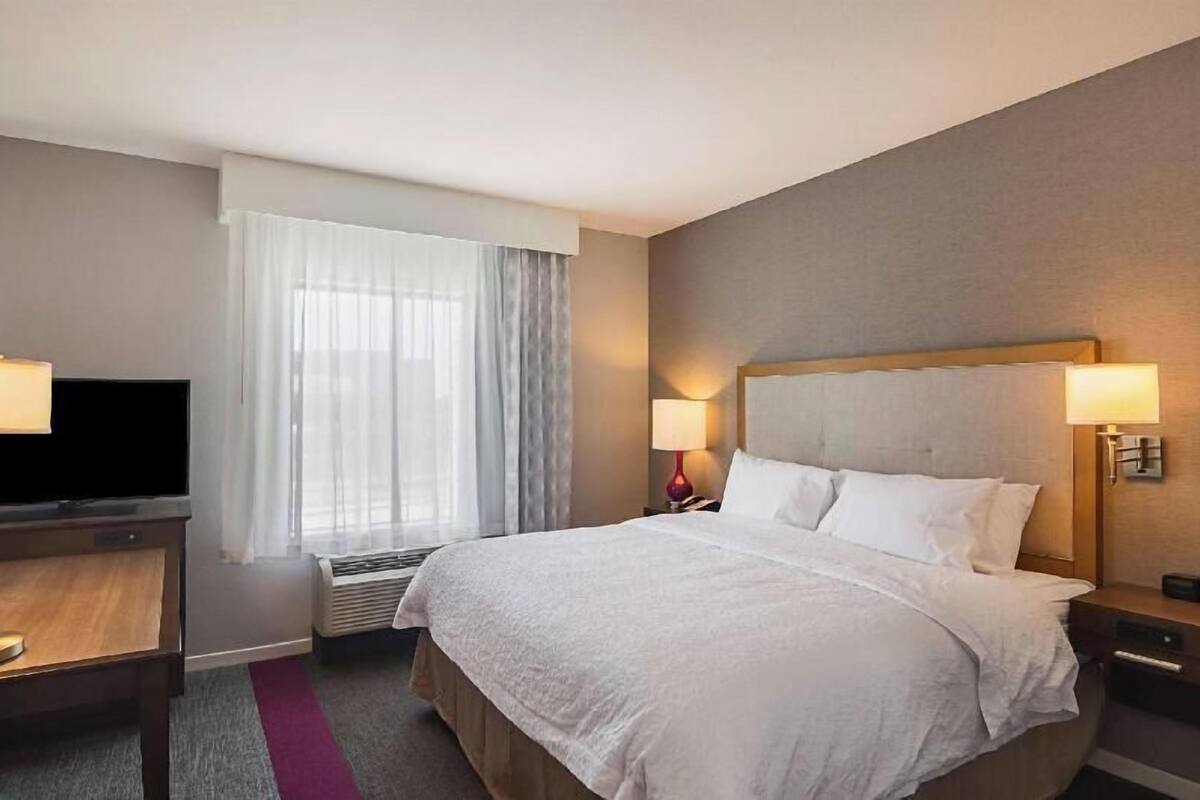 Habitación de hotel con cama, lámparas de noche, televisor, ventana con cortinas y un calentador fijado a la pared.
