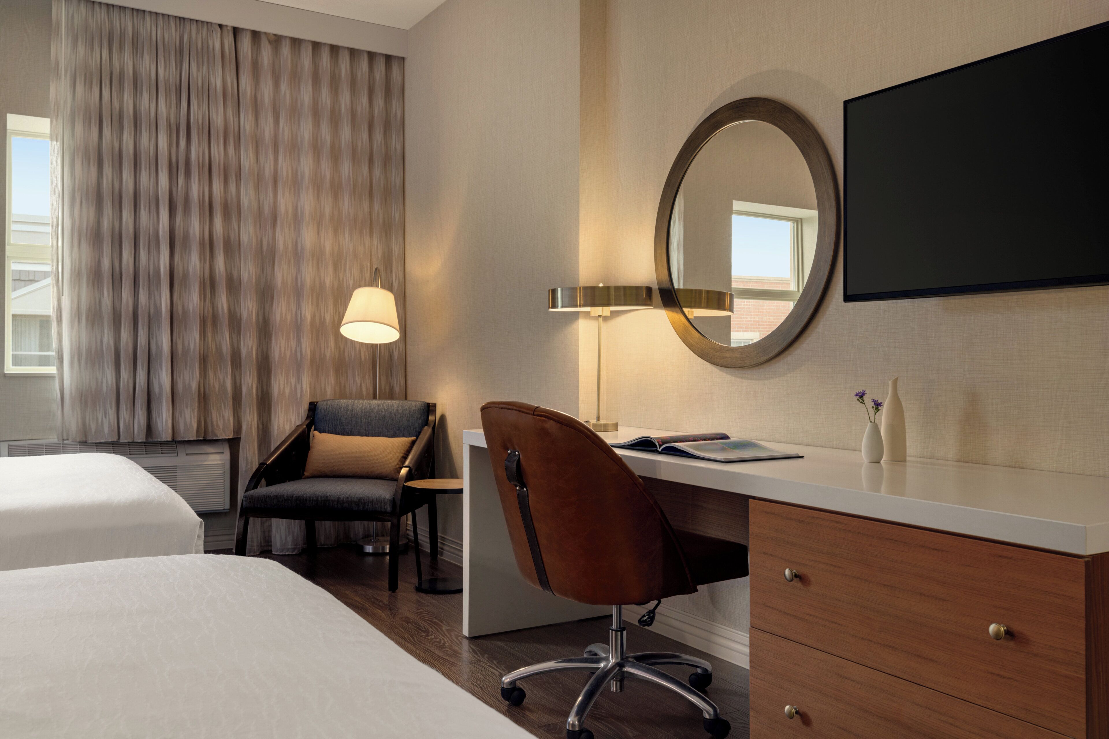 A hotel room with two beds, a desk, a chair, a lamp, a mirror, and a television.