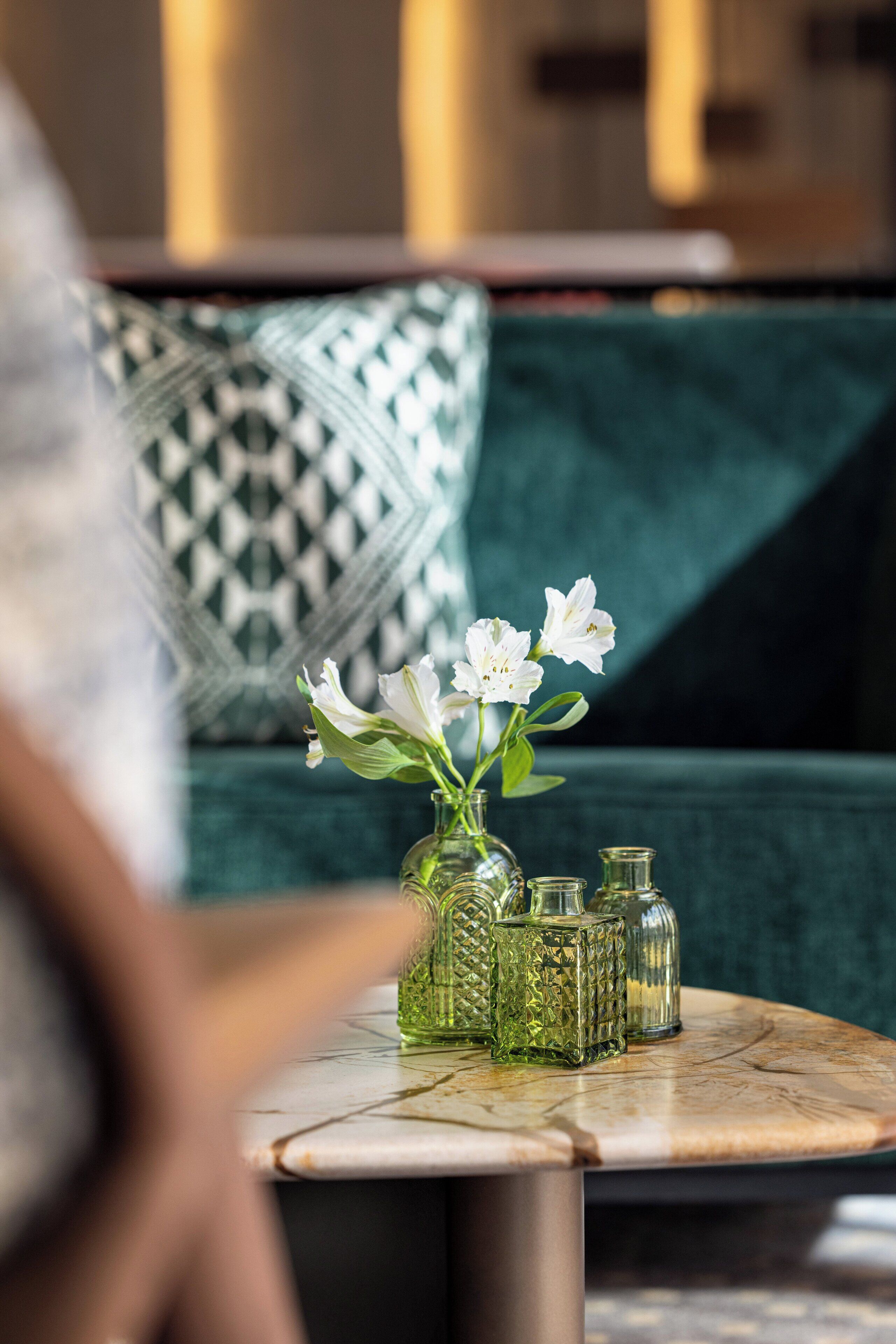A green sofa with a patterned cushion, a small round table with a vase of white flowers, and several glass bottles.