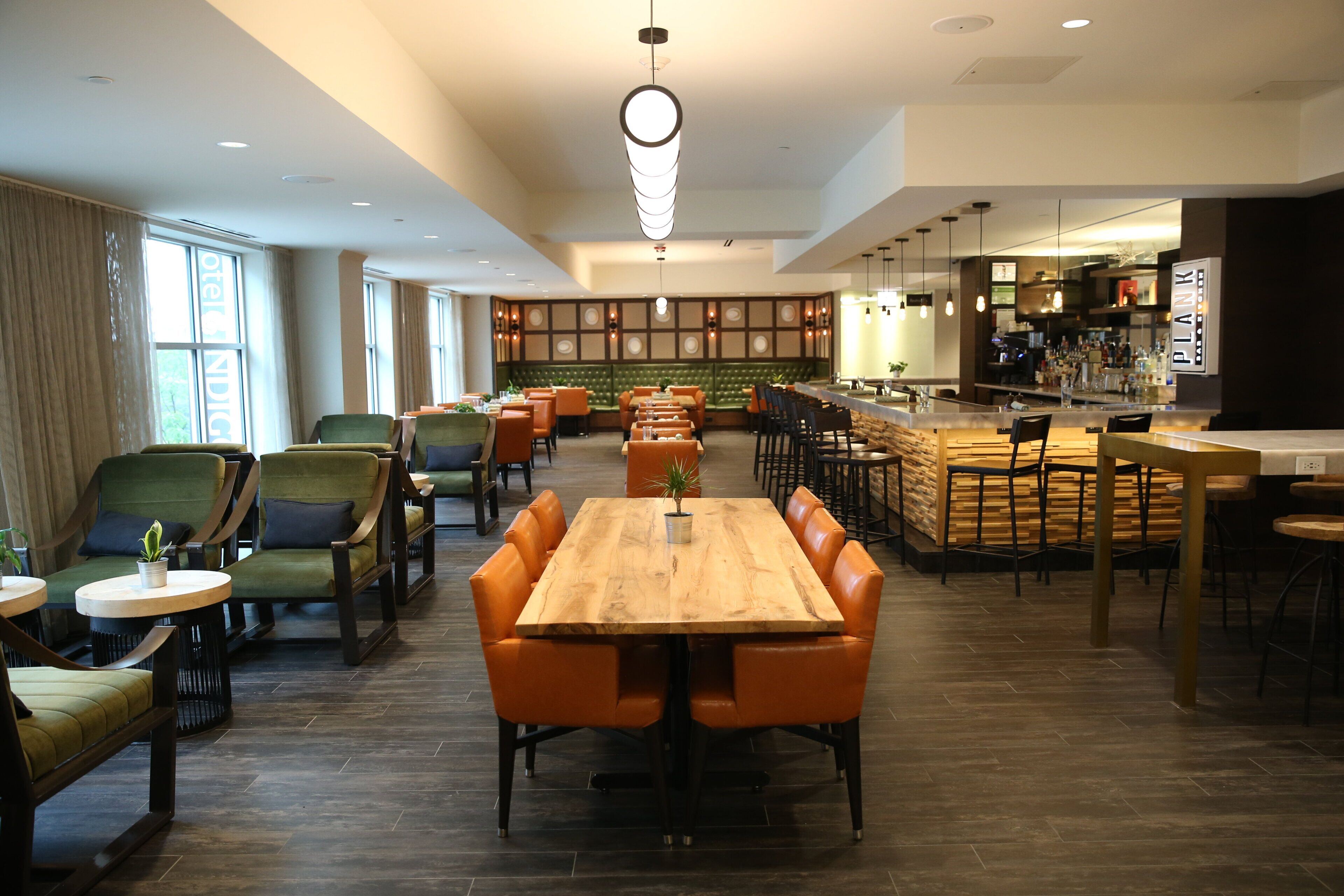 Un restaurante moderno con una mesa de madera central rodeada de sillas naranjas, una zona de bar y un área de estar con sofás verdes tapizados.