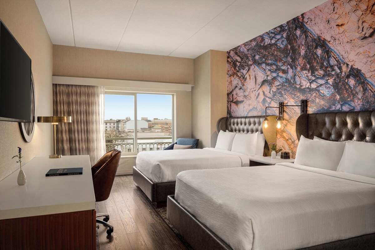 Une chambre d’hôtel avec deux lits, un bureau, une chaise, une télévision et une grande fenêtre donnant sur la ville.