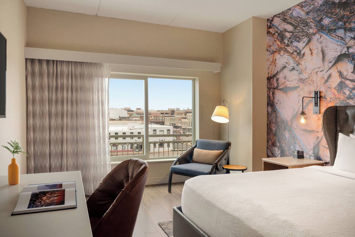 Une chambre d’hôtel avec un grand lit, une chaise, une petite table, une lampe et une vue sur la ville par la fenêtre.