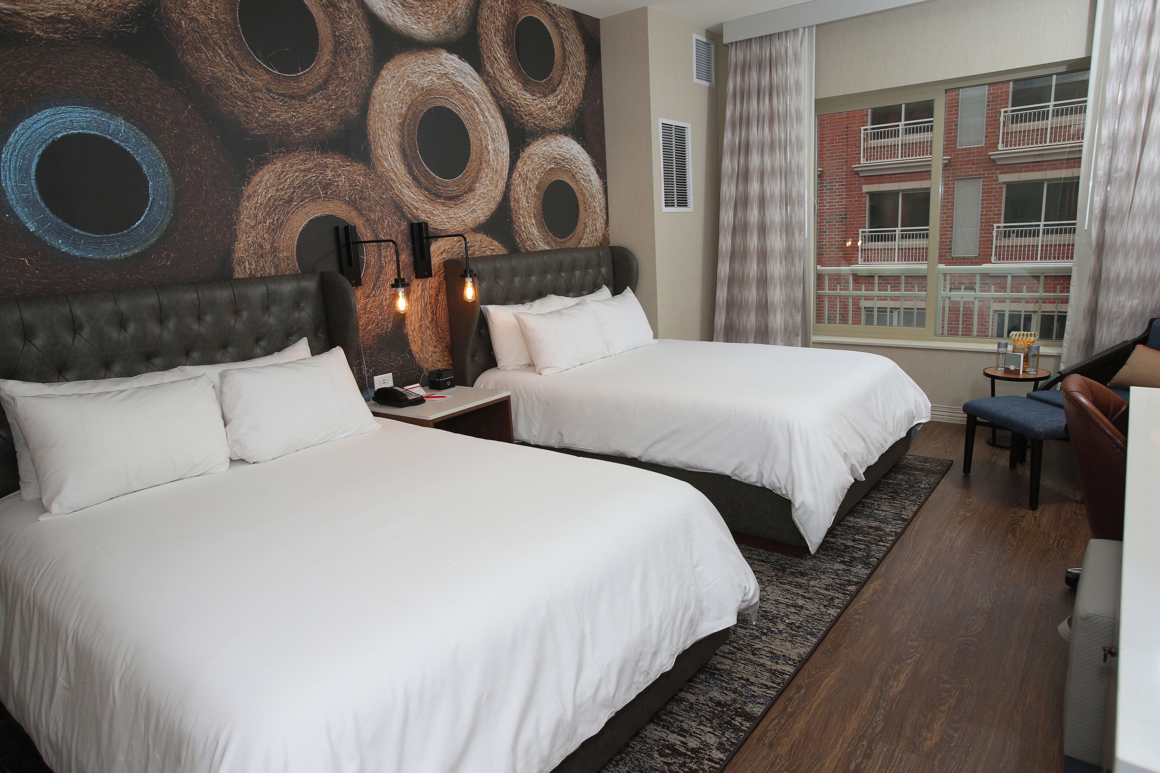 Habitación de hotel con dos camas, un mural grande en la pared, una ventana con vista a un edificio y una pequeña mesa con una silla.