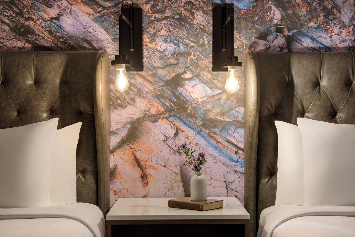 Une chambre d’hôtel avec une tête de lit capitonnée, une table de chevet ornée d’un vase et d’un livre, et un mur décoré d’un motif imitant le marbre.