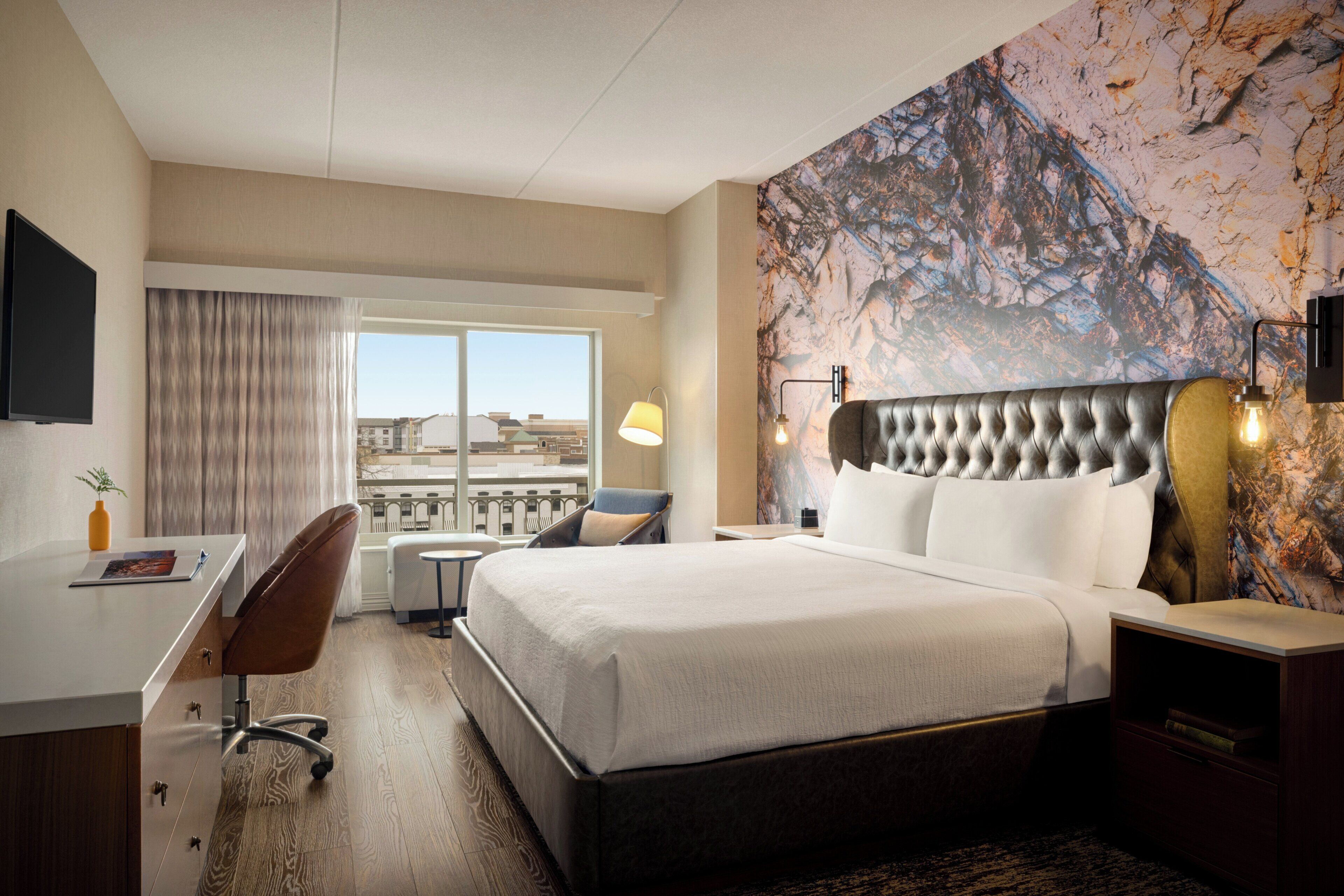 A hotel room with a large bed, a desk, a chair, a desk lamp, and a view of the city through the window.