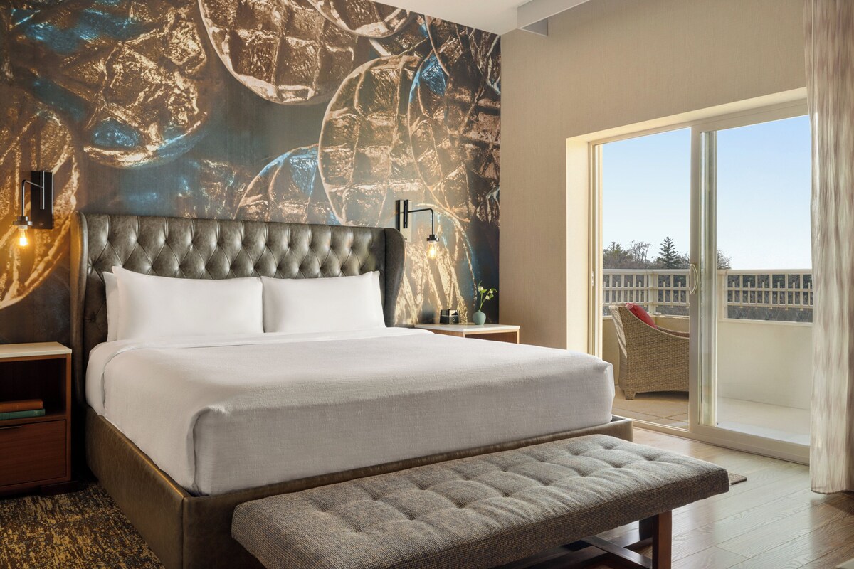 Une chambre moderne avec un grand lit, un banc, une vue sur l’extérieur et un mur décoré d’œuvres d’art abstraites.