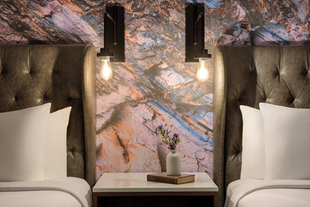 Une pièce avec un mur décoré d’un motif imitant le marbre, deux têtes de lit capitonnées, une table de chevet ornée d’un vase et d’un livre, et deux appliques murales.