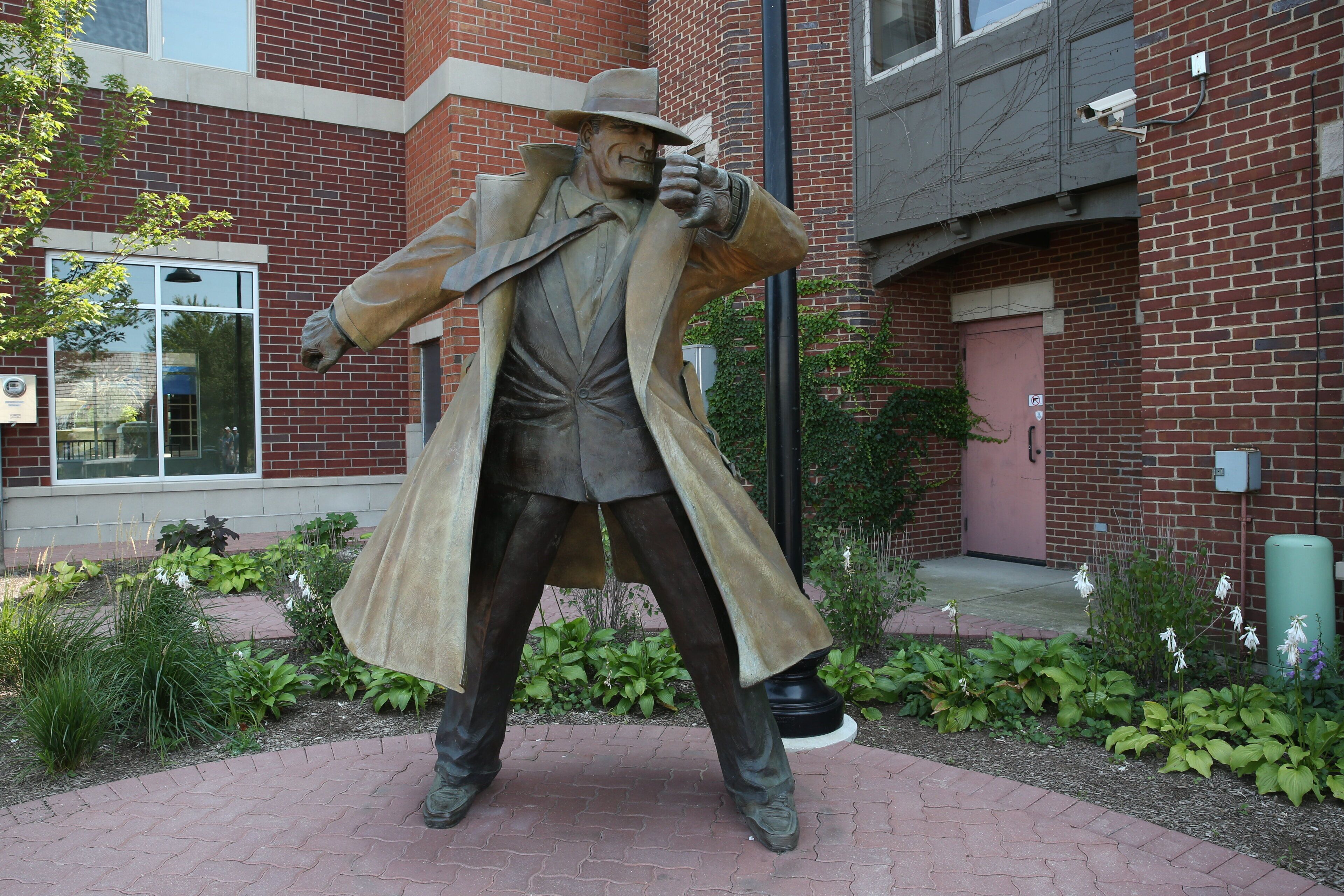 A statue of a man in a trench coat and hat, positioned in front of a brick building with a window and a door.