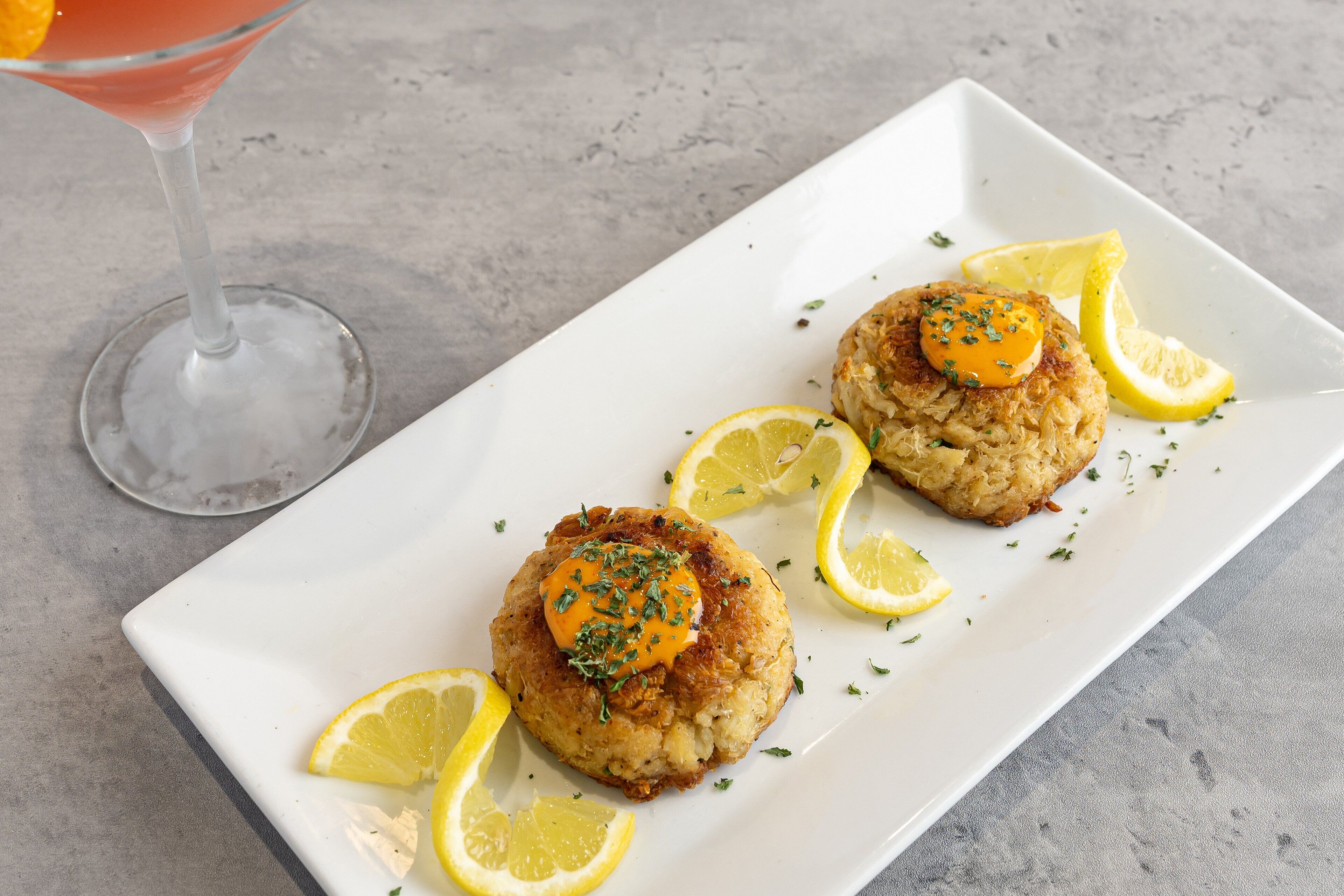 A white rectangular plate with two crab cakes topped with a dollop of sauce and garnished with herbs, accompanied by lemon slices.