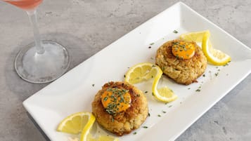 A white rectangular plate with two crab cakes topped with a dollop of sauce and garnished with herbs, accompanied by lemon slices.