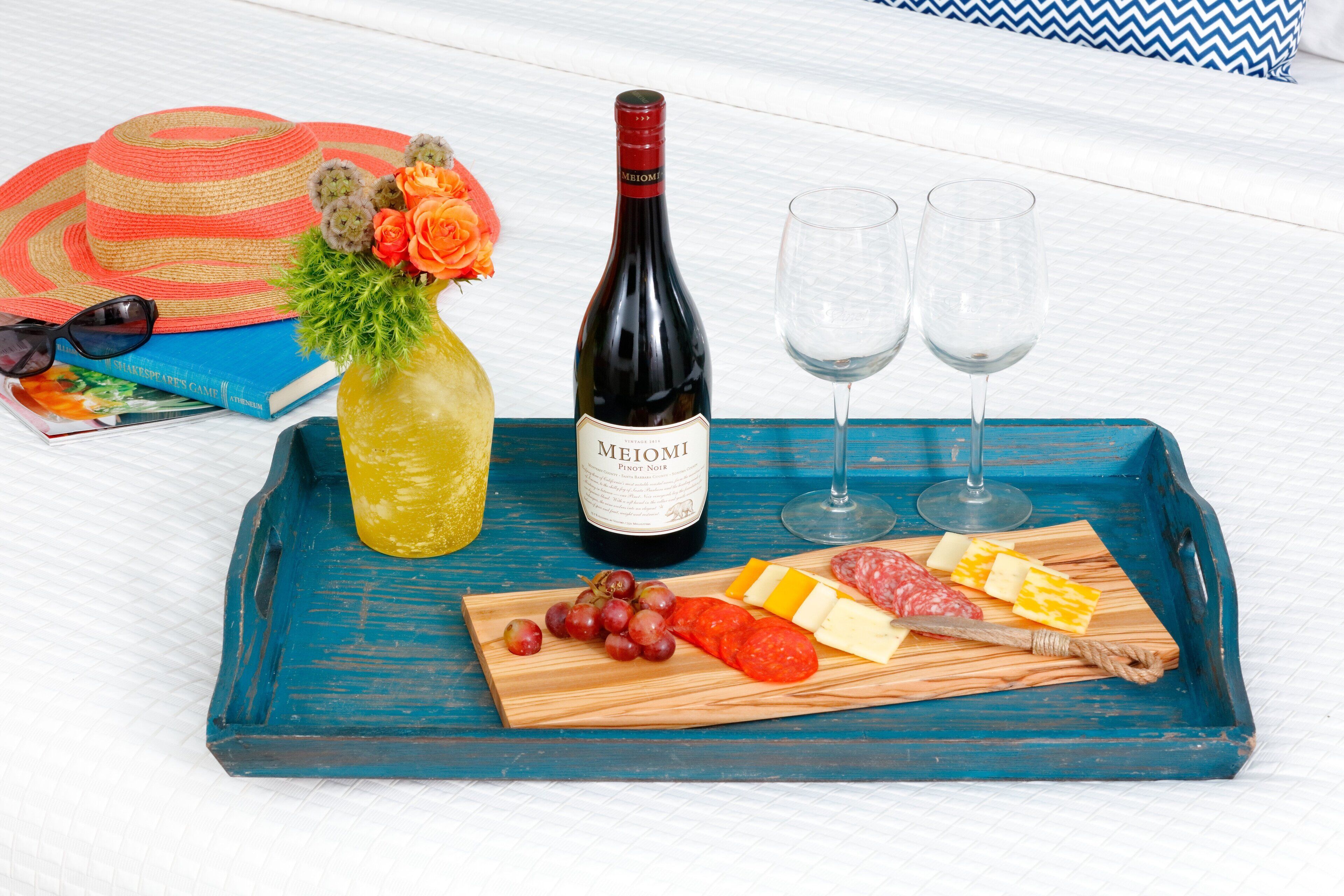 Un plateau contenant une bouteille de vin Meomi, deux verres, un chapeau, un livre, un bouquet de fleurs, ainsi qu’un assortiment de fromages et de charcuteries.