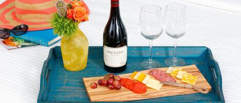 Un plateau contenant une bouteille de vin Meomi, deux verres, un chapeau, un livre, un bouquet de fleurs, ainsi qu’un assortiment de fromages et de charcuteries.