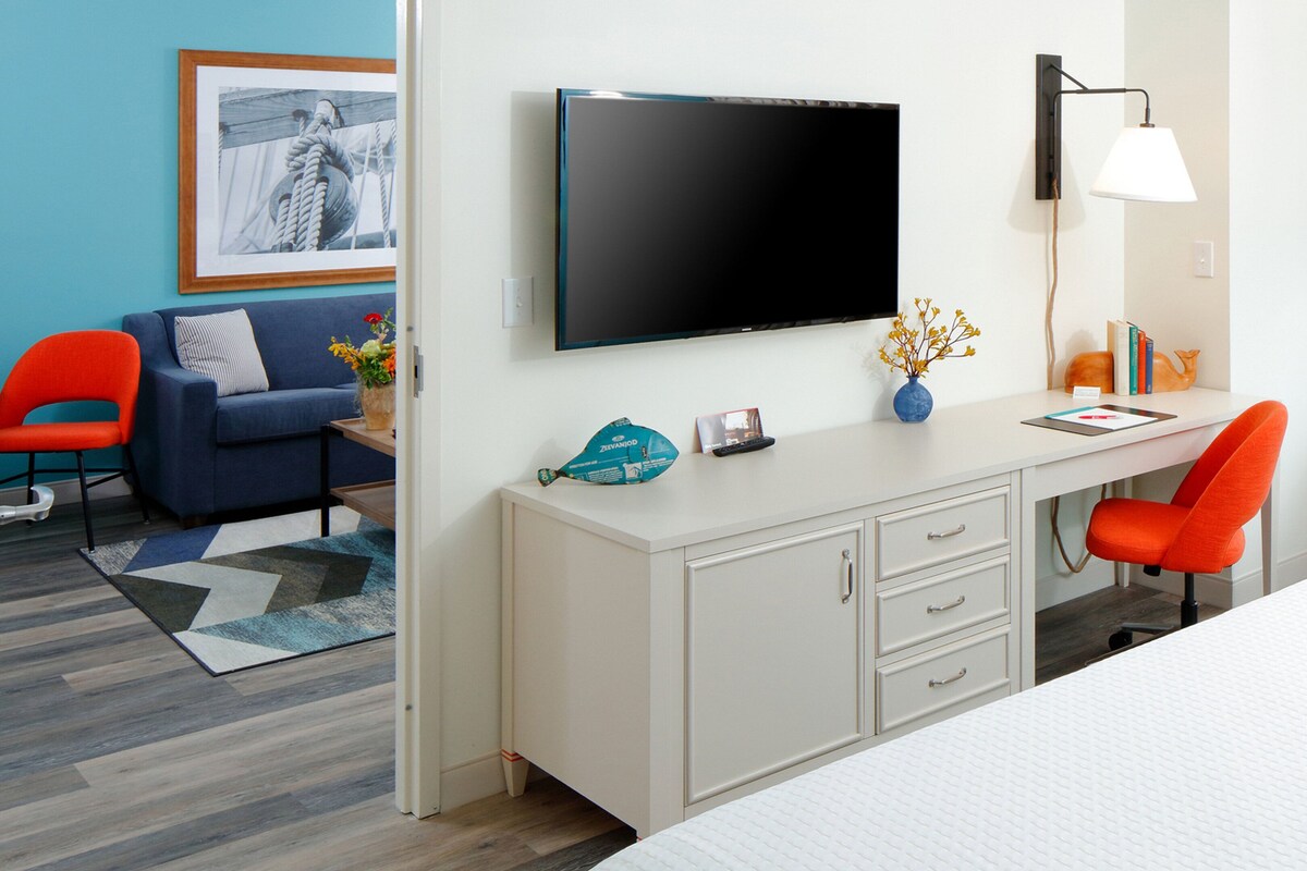 Habitación de hotel con cama, escritorio con silla, televisor, ventana con vistas y un cuadro en la pared.