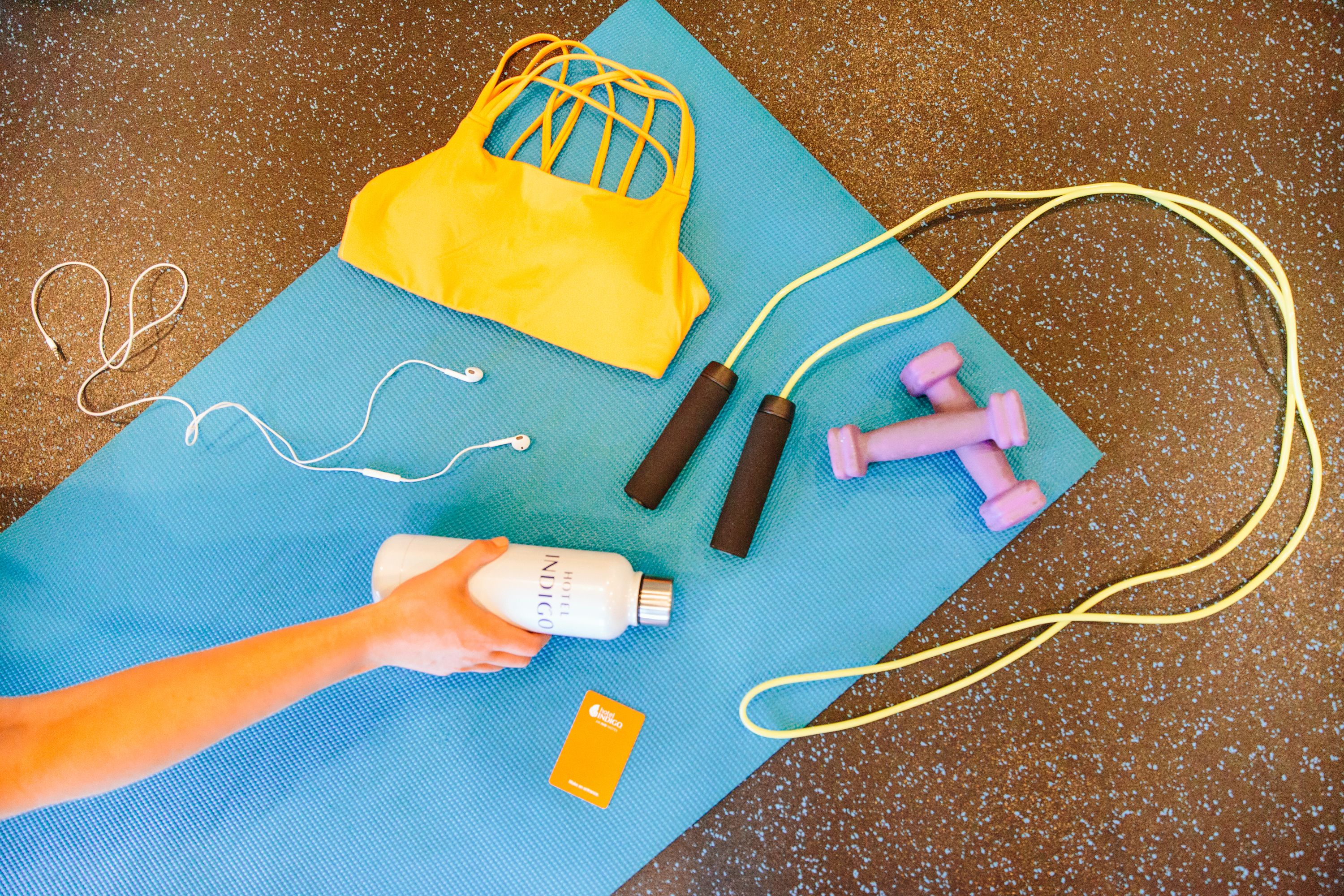 A blue exercise mat with a yellow bag, pink dumbbells, a jump rope, and a bottle of lotion.