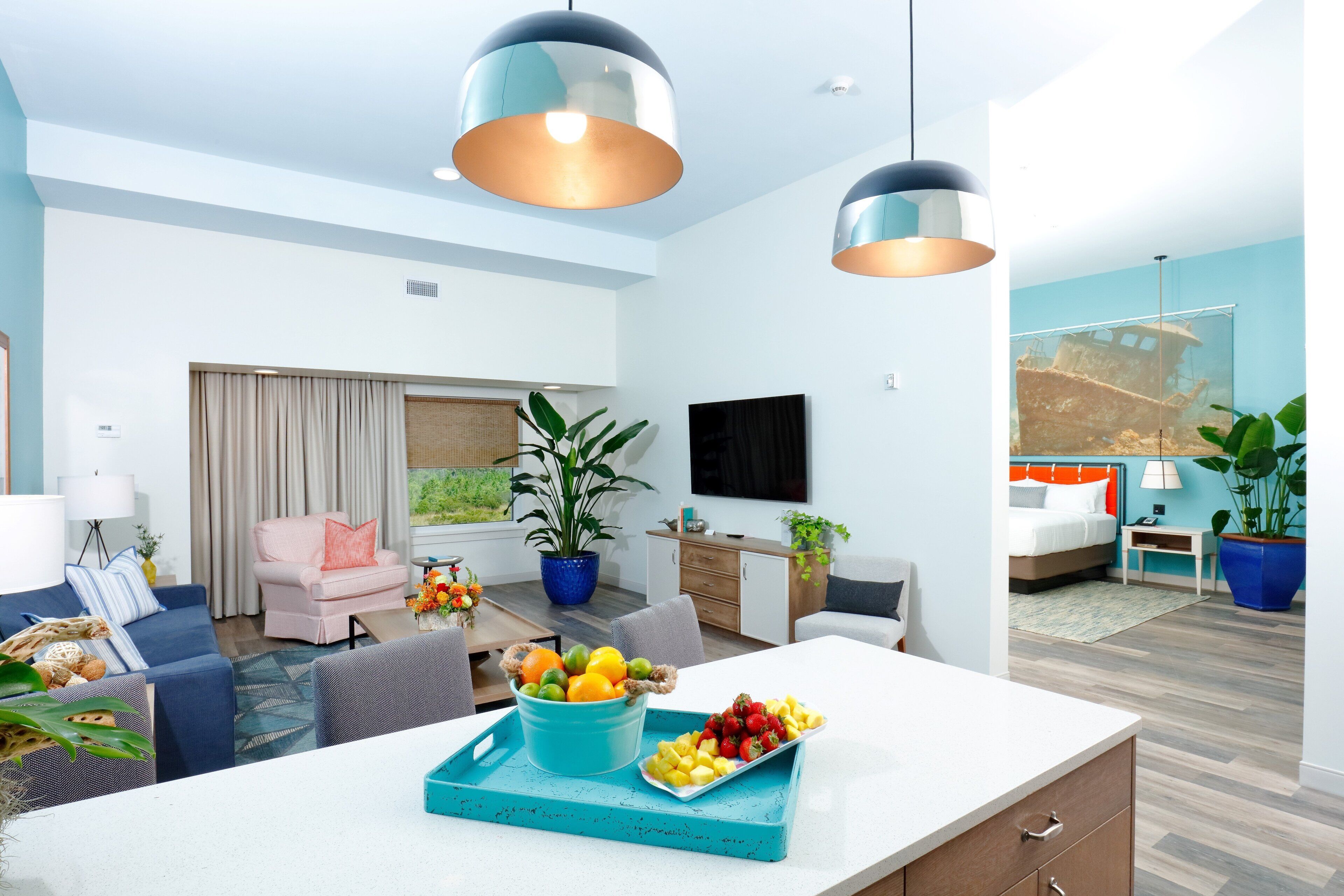 A modern living room with a sofa, a dining table with fruit, a TV, and a large window.