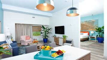 A modern living room with a sofa, a dining table with fruit, a TV, and a large window.