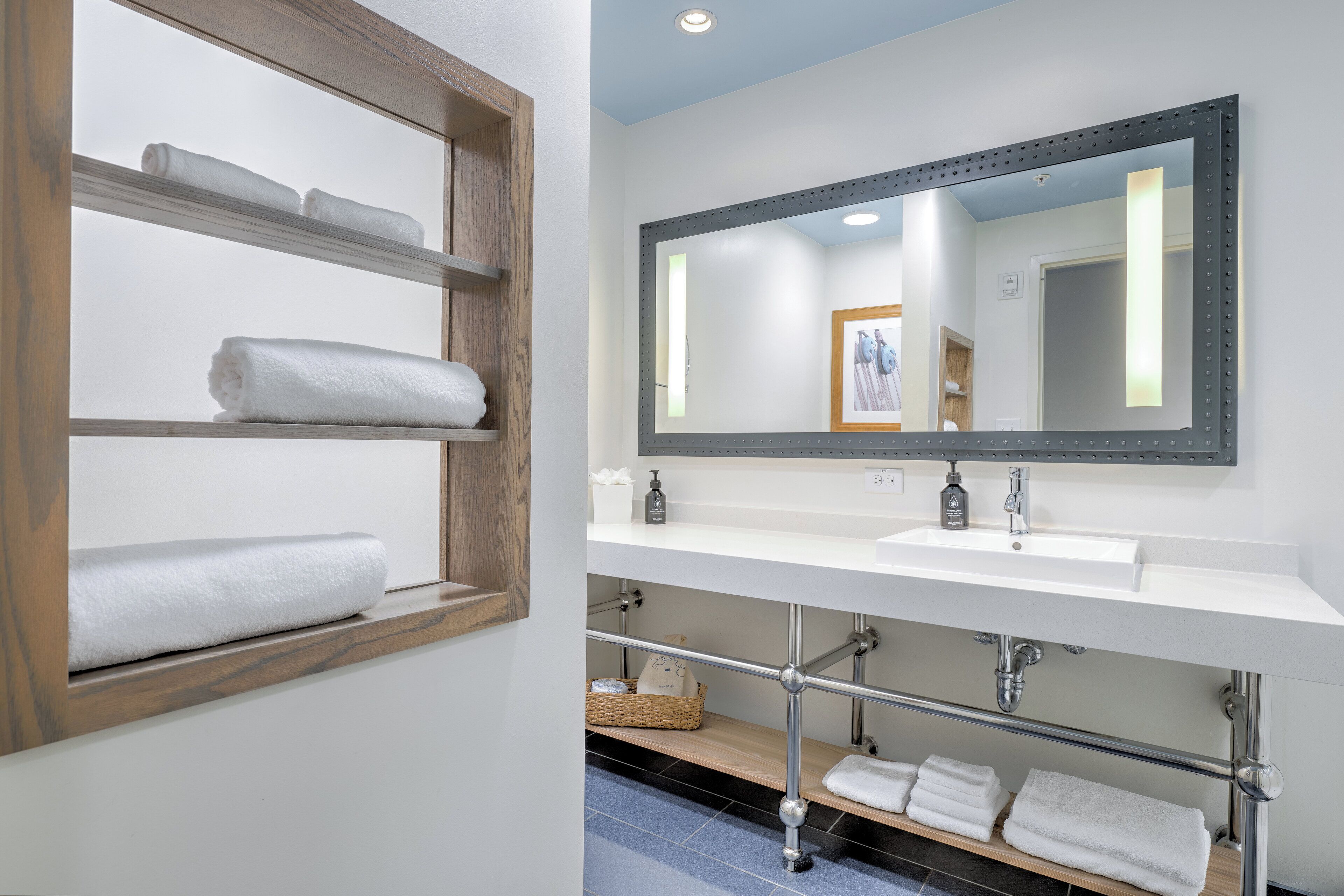 A bathroom with a wooden shelf holding towels, a white sink with a mirror above it, and a tiled floor.