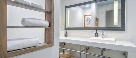 A bathroom with a wooden shelf holding towels, a white sink with a mirror above it, and a tiled floor.