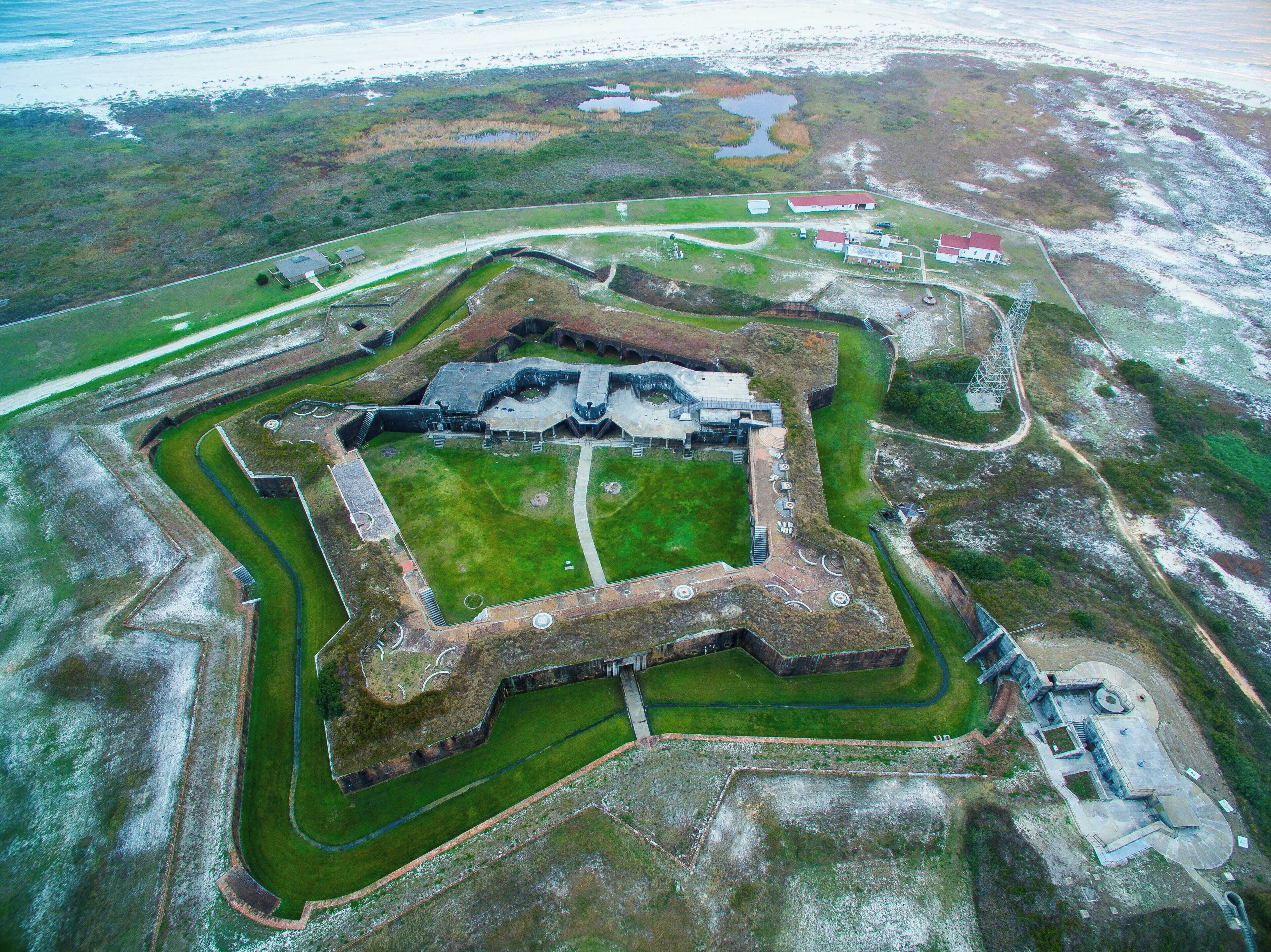 Vue aérienne d’une structure fortifiée comprenant un bâtiment central, des murs d’enceinte et un fossé.