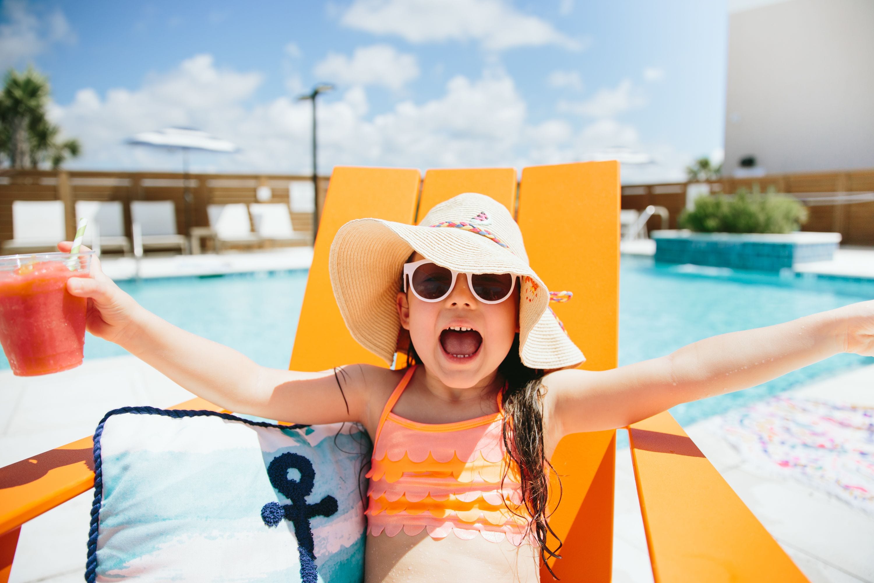 Un enfant assis sur une chaise au bord de la piscine, les bras tendus, tenant une boisson.