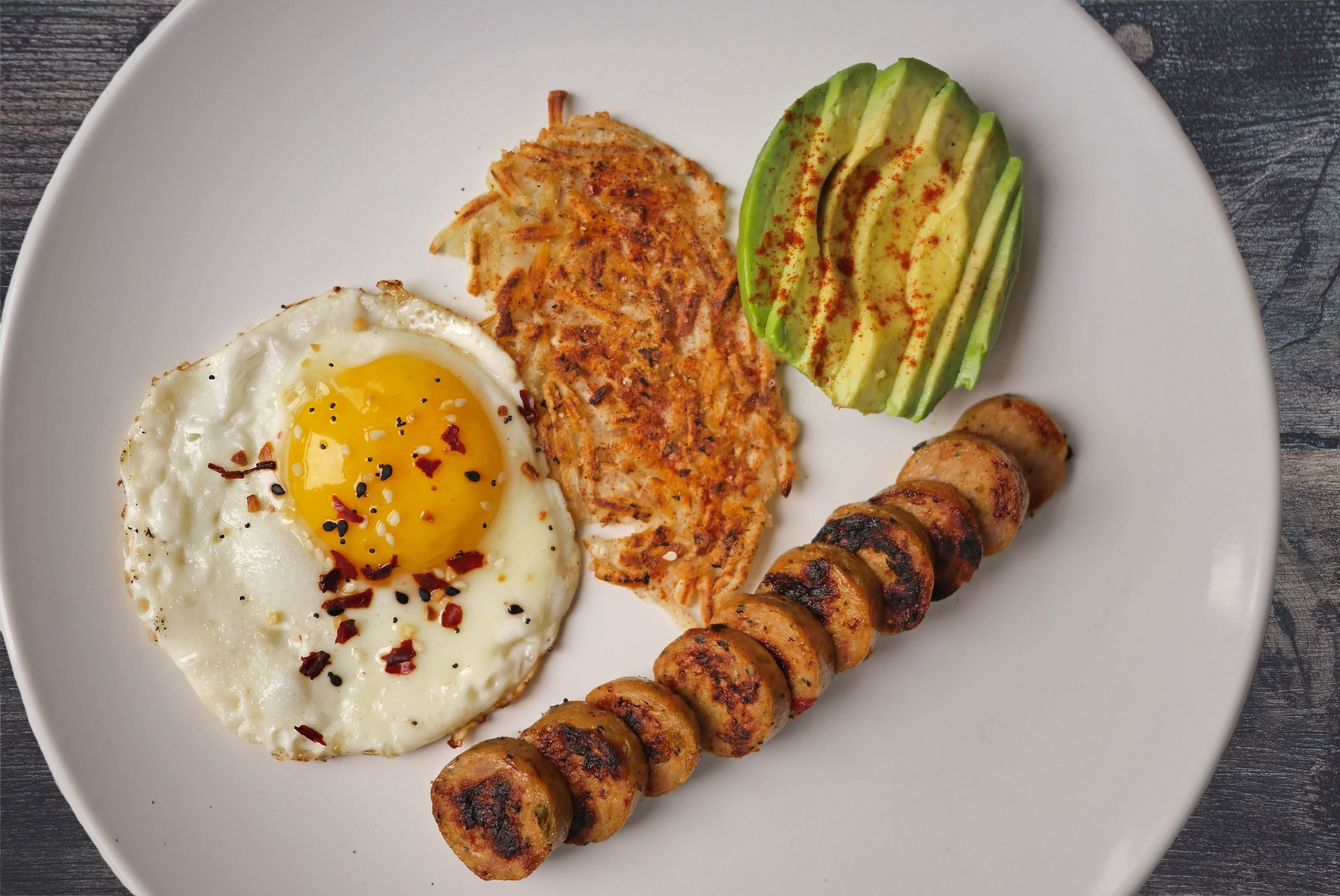 A plate with a fried egg, hash browns, grilled sausage, and sliced avocado.
