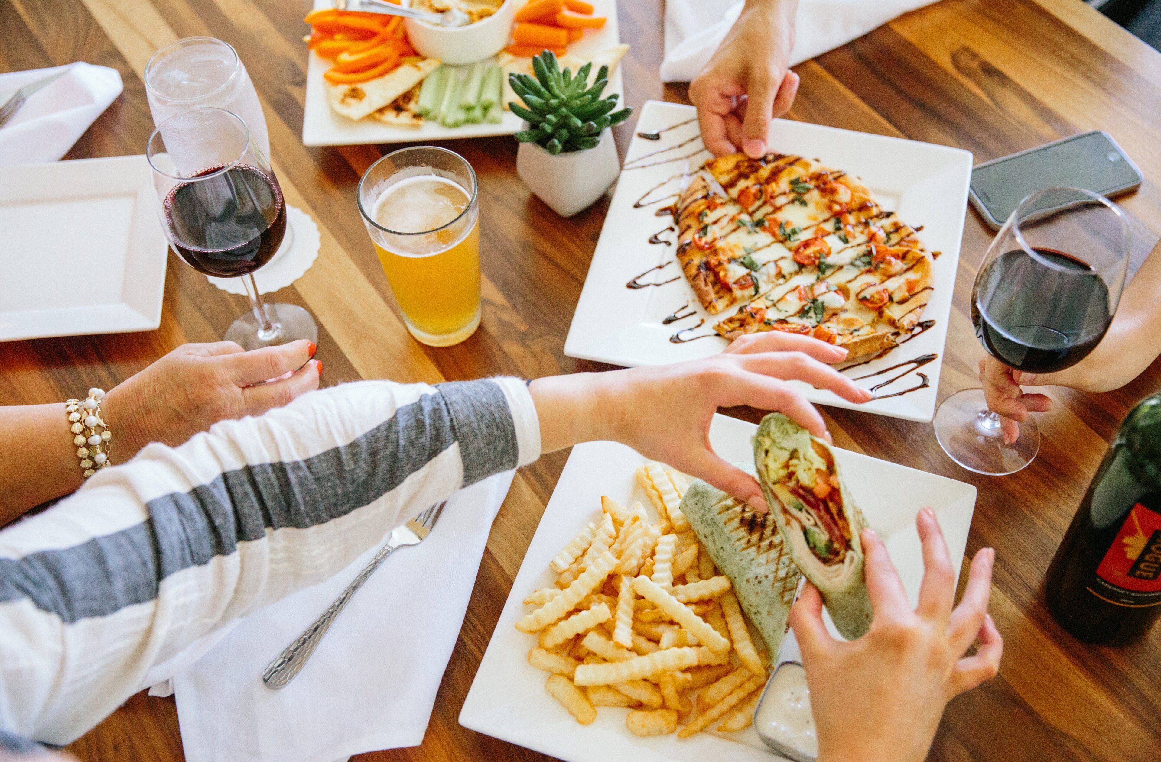 A group of people dining at a wooden table with various dishes, drinks, and a smartphone.