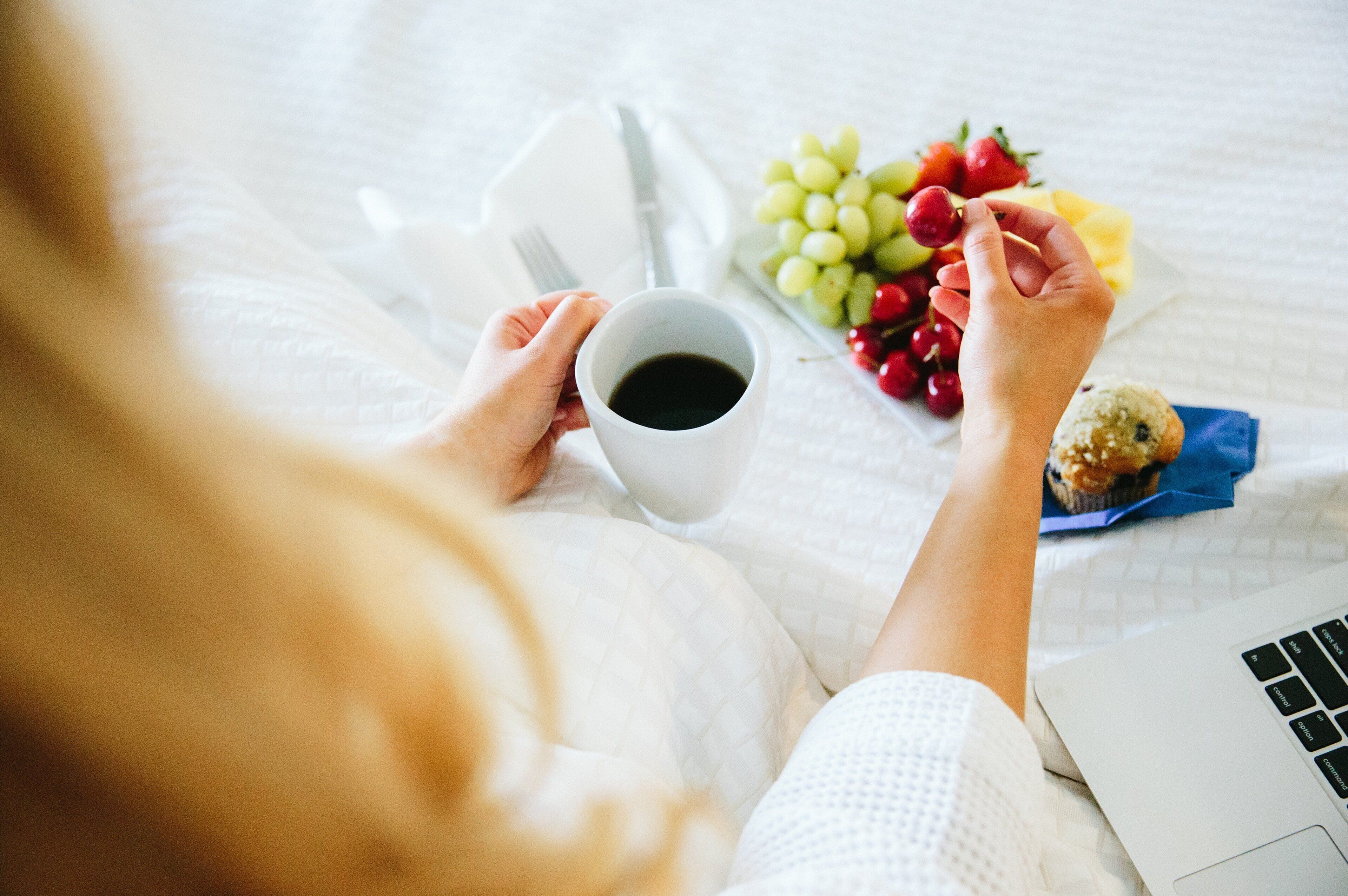 A person holding a cup of coffee, a plate of fruit, and a muffin on a bed with a laptop.