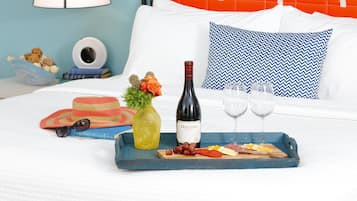 Uma bandeja de café da manhã bem organizada com uma garrafa de vinho Meomi, copos, uvas, queijo e um chapéu sobre uma cama com travesseiro azul e branco, uma luminária e um relógio de parede.