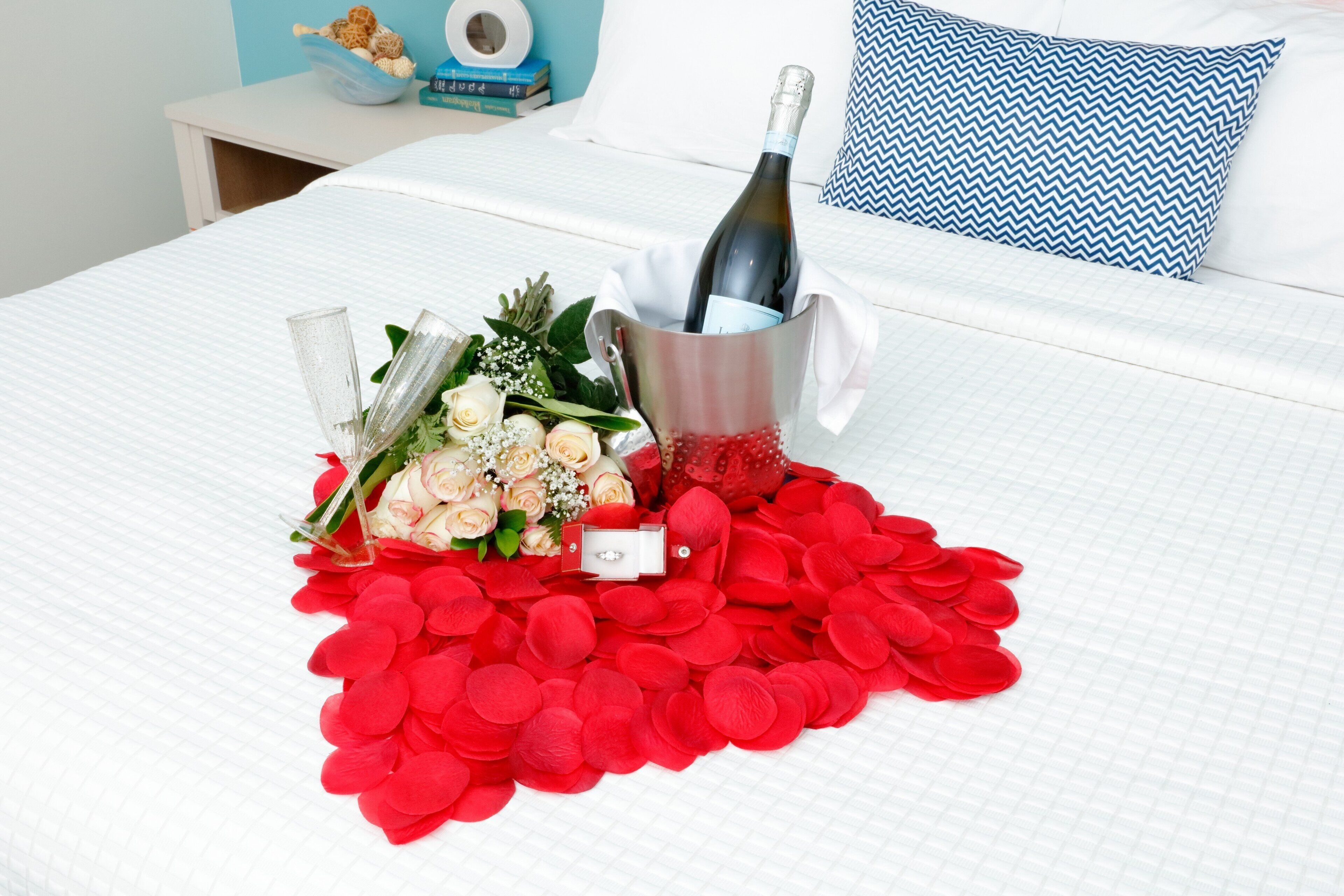 A bed with a bouquet of flowers, a bottle of champagne, and red rose petals.