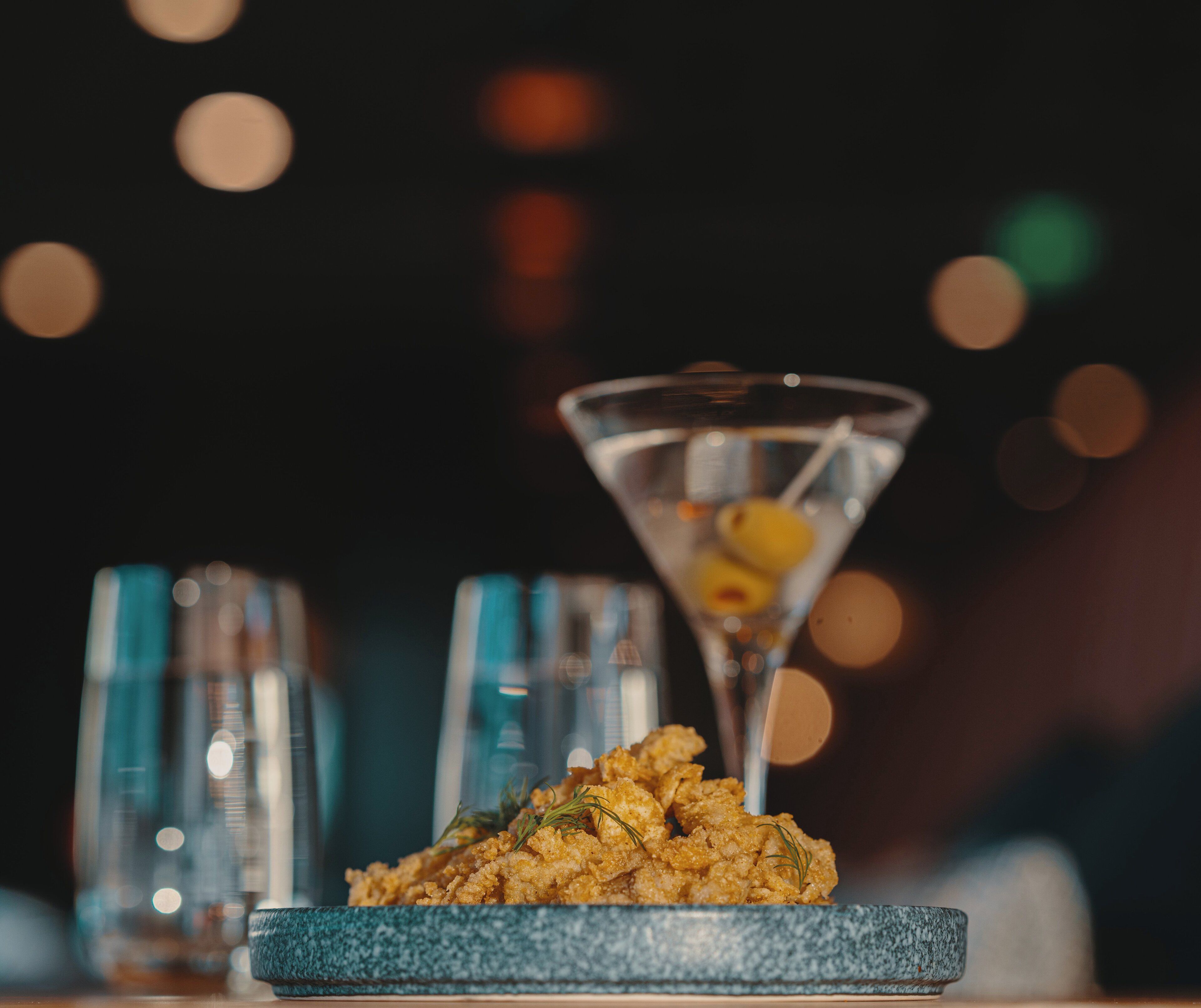 A martini glass with a lemon slice on a blue plate, accompanied by a glass of water and a bottle of olive oil.