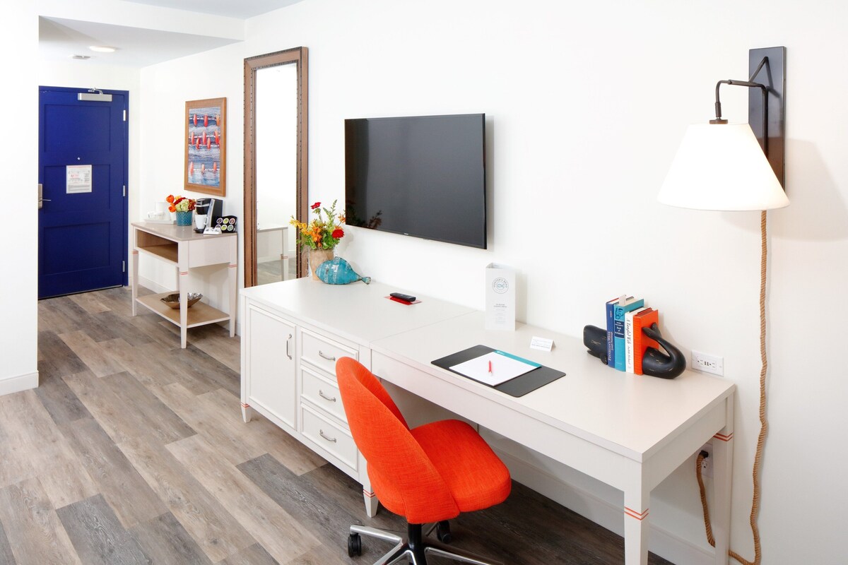 A modern hotel room with a white desk, a red chair, a flat-screen TV, a lamp, and a blue door.