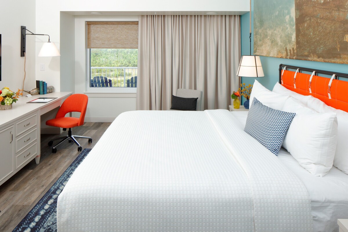 A hotel room with a large bed, a desk with a chair, a window with curtains, and a painting on the wall.